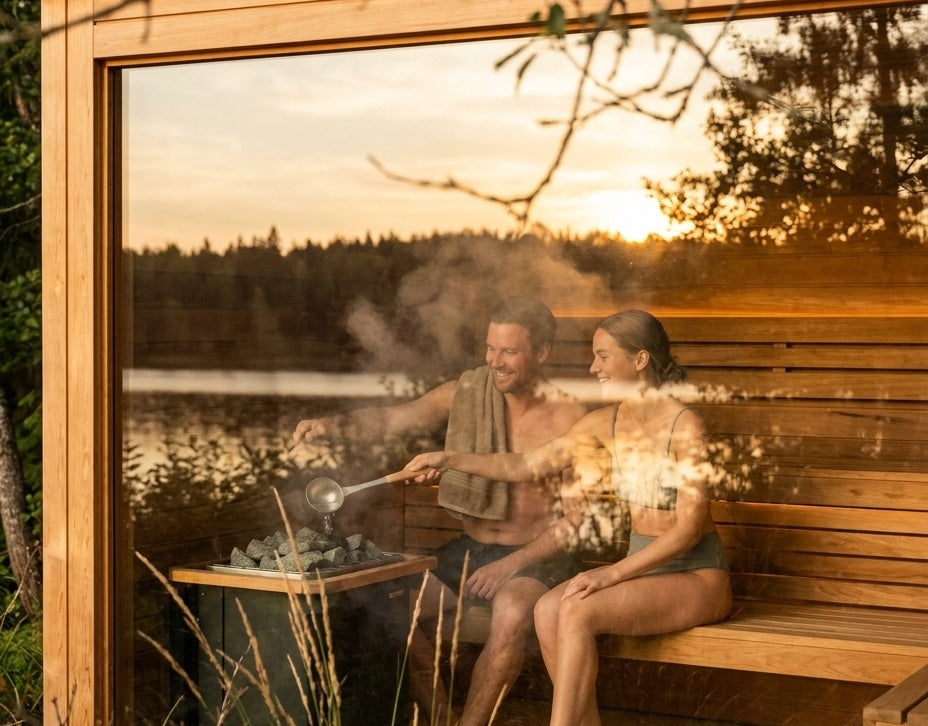 A couple sitting in a sauna looking outside through a glass front on a lake at sunset
