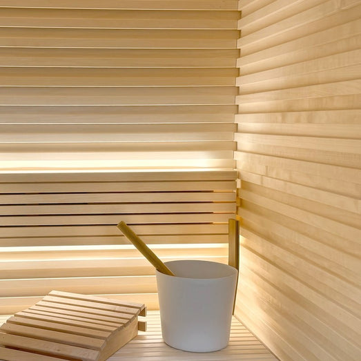 Sauna interior with horizontal slatted wall paneling, a bench, a white bucket with ladle and a wooden headrest