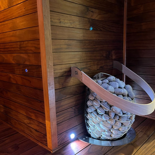 Sauna interior with horizontal wall paneling and a heater basket filled with stones inside a curved wooden rail