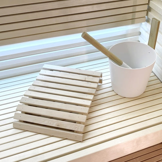Sauna bench with a slatted wooden headrest, a white bucket with a wooden ladle, and horizontal slatted wall paneling