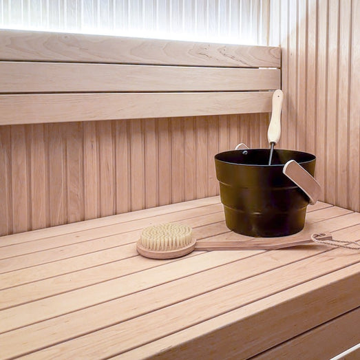 Sauna interior with vertical wall paneling, tiered benches, a dark bucket with ladle and a long-handled brush