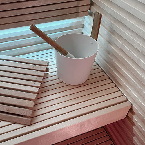 Sauna interior with slatted benches, a white bucket with wooden ladle, and horizontal wall paneling with blue backlight