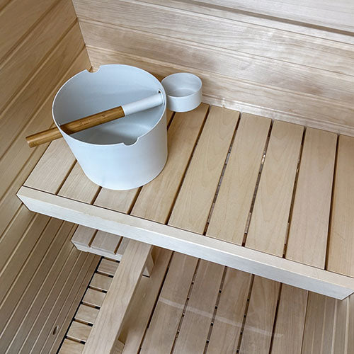 Sauna interior with slatted two-tier benches, paneled walls, and a white bucket with a wooden ladle on the upper bench