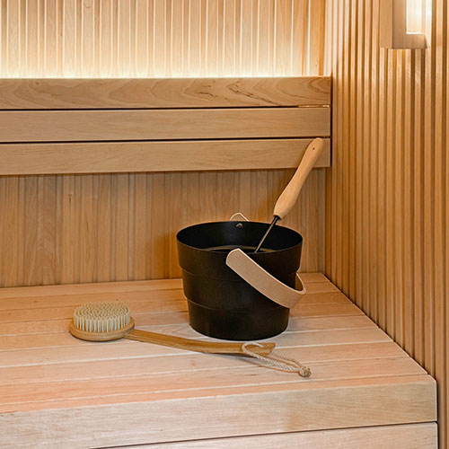 Sauna interior with slatted wall paneling, tiered benches, a dark bucket with ladle and a long-handled brush
