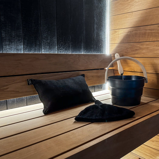Sauna bench with a black pillow, sauna hat and bucket, horizontal paneling and a dark accent wall with LED light