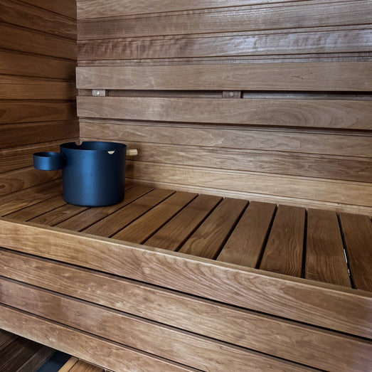 Slatted sauna bench with a dark bucket in the corner and horizontal wall paneling behind it