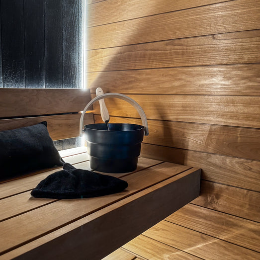 Sauna bench with a black pillow, sauna hat and bucket against horizontal wall paneling with a lit edge