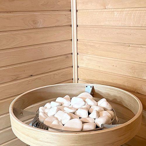 Round wooden heater rim holding white stones in a corner of a sauna with horizontal wall paneling