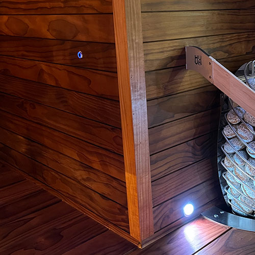 Sauna interior corner with horizontal wood paneling, a small wall light, and a heater with stones behind a rail