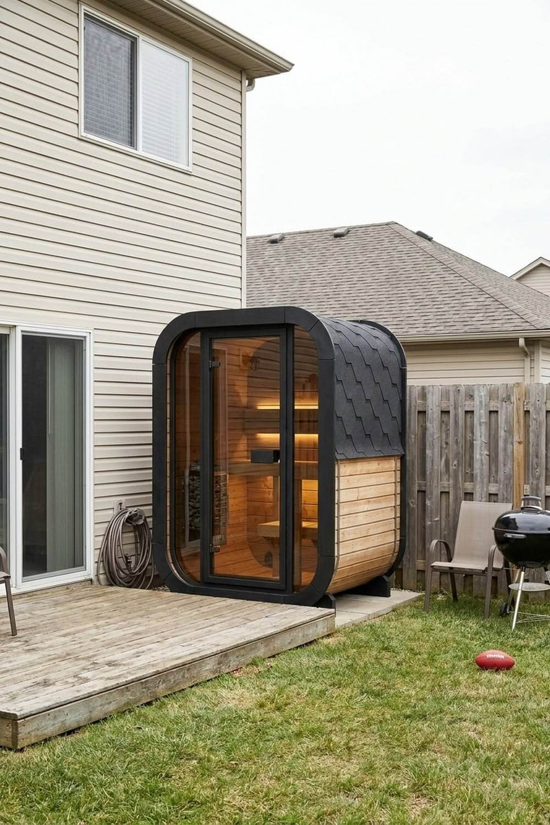 SaunaLife CL3G cube sauna with a black frame and glass front, glowing warm beside the wooden deck of an ordinary backyard, with a fence, a grill and a football on the lawn