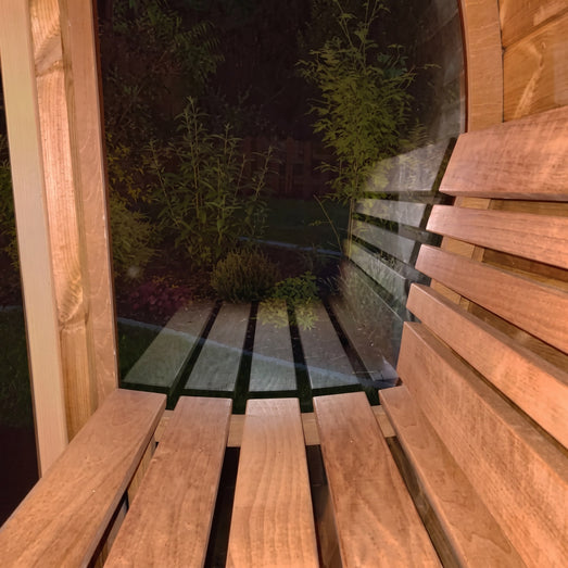 SaunaLife E8 barrel sauna bench beside window at night overlooking lit garden