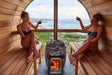 Two people relaxing in a barrel sauna with a HUUM wood stove and ocean view