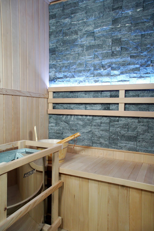 Interior of the SunRay Westlake 3-person indoor traditional sauna with slate-colored stone back wall, lit backrest, bench and Harvia heater in a wooden guard
