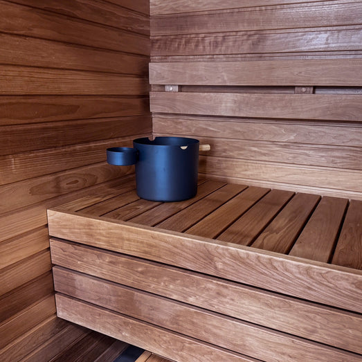 Corner of a sauna with horizontal wall paneling, a slatted bench and a dark bucket